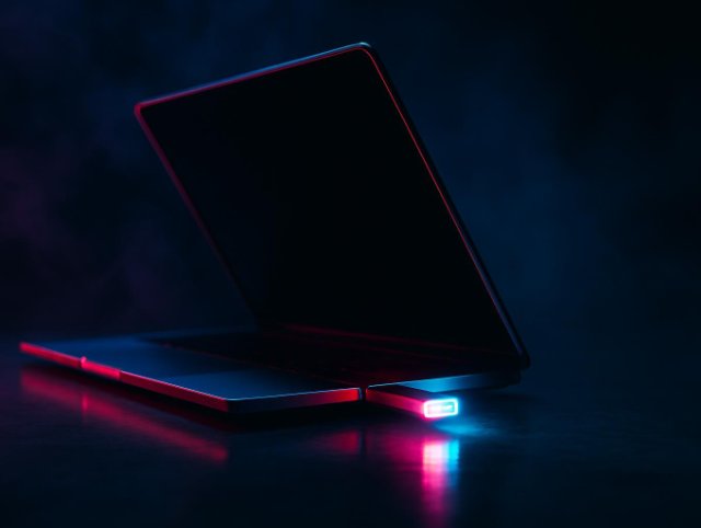 USB flash drive plugged into the side port of a half-open laptop, glowing in pink and blue neon studio lighting